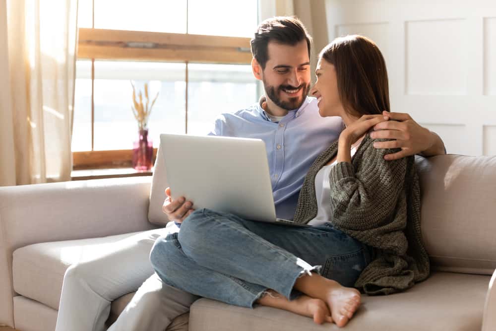 A man and woman sit while on a laptop, looking at one another on the couch.