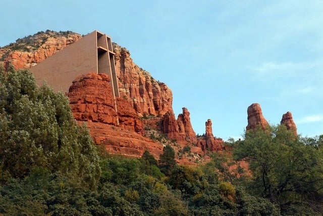 A large building on the side of a red rock formation with green trees along the ground below it.