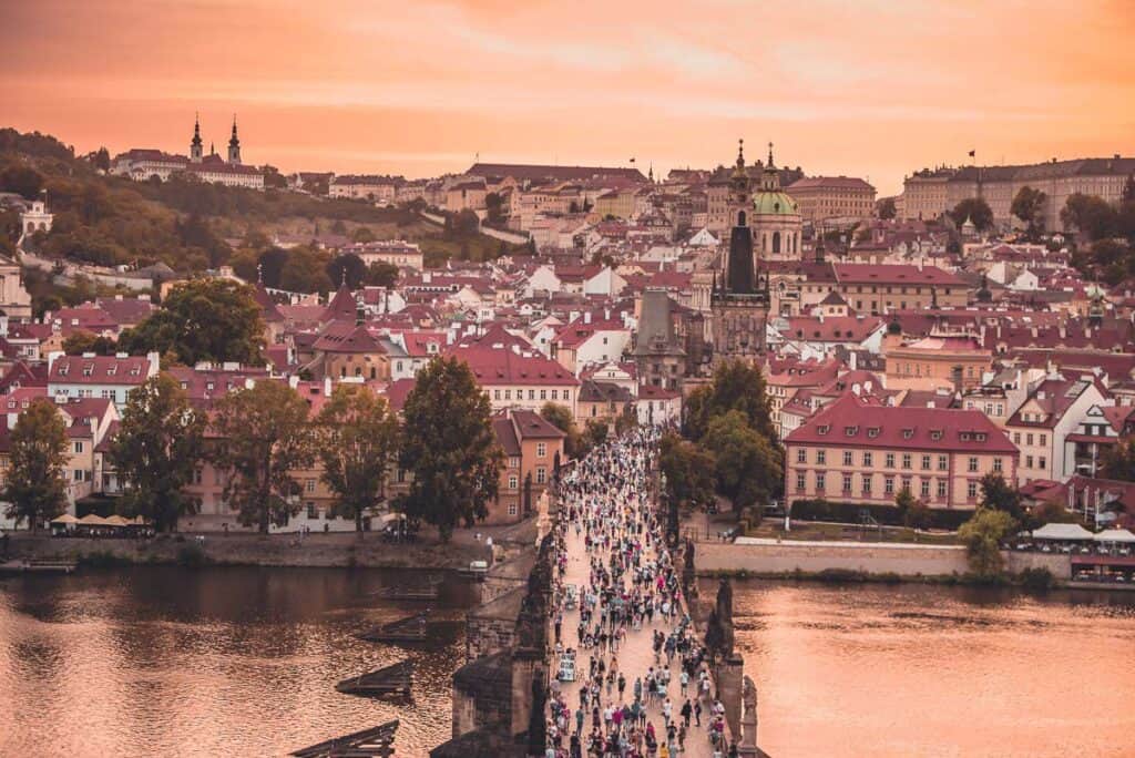 A crowd of people walking across a bridge over a river leading to a European town.